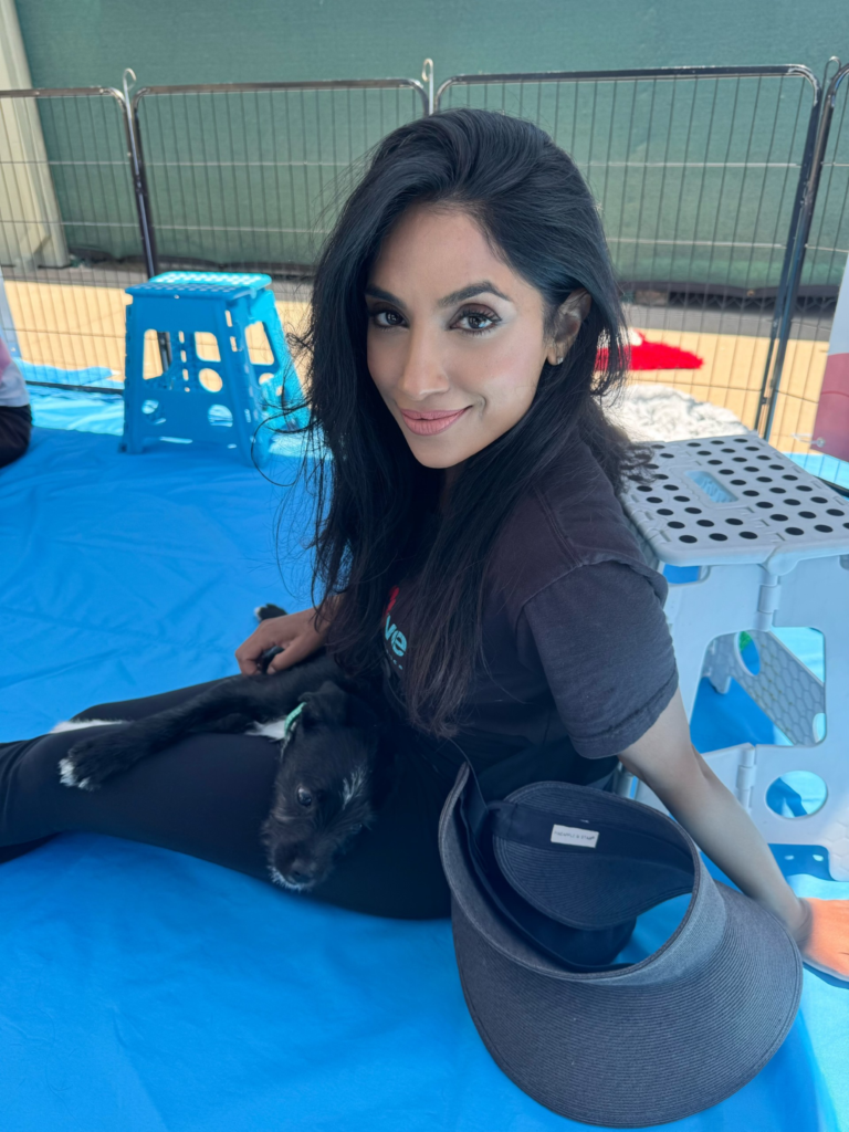 Woman named sony mathew with long dark hair, wearing a black shirt, smiling while sitting with a small black puppy at an outdoor event in los angeles